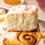 A frosted cinnamon roll resting on a spatula, hovering over a baking dish of freshly baked cinnamon rolls.