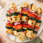 Grilled chicken kabobs on a paper plate with red onion, bell pepper, and zucchini.