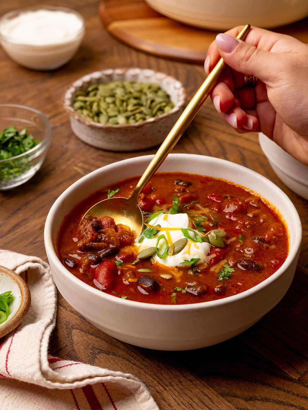 A hand with a spoon in a white bowl of chili surrounded by ingredients.