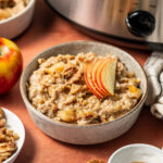 A bowl of apple cinnamon oatmeal in front of a slow cooker topped with apple slices.