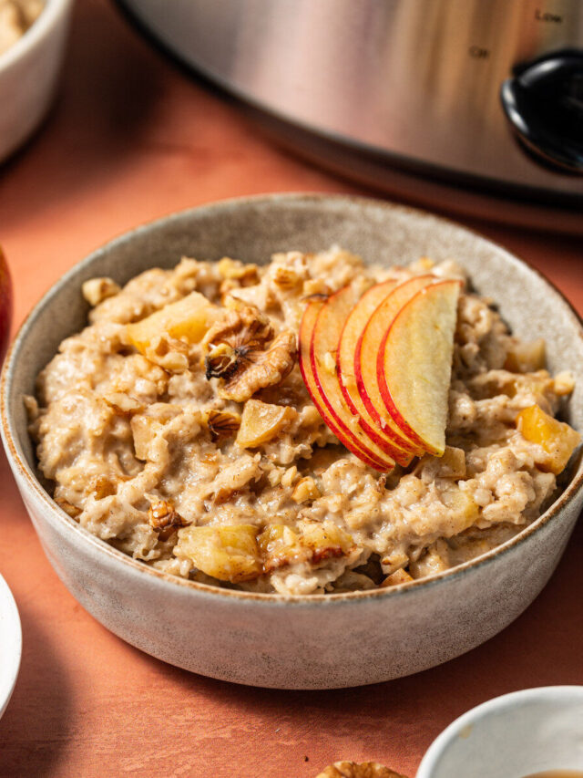 A bowl of apple cinnamon oatmeal in front of a slow cooker topped with apple slices.