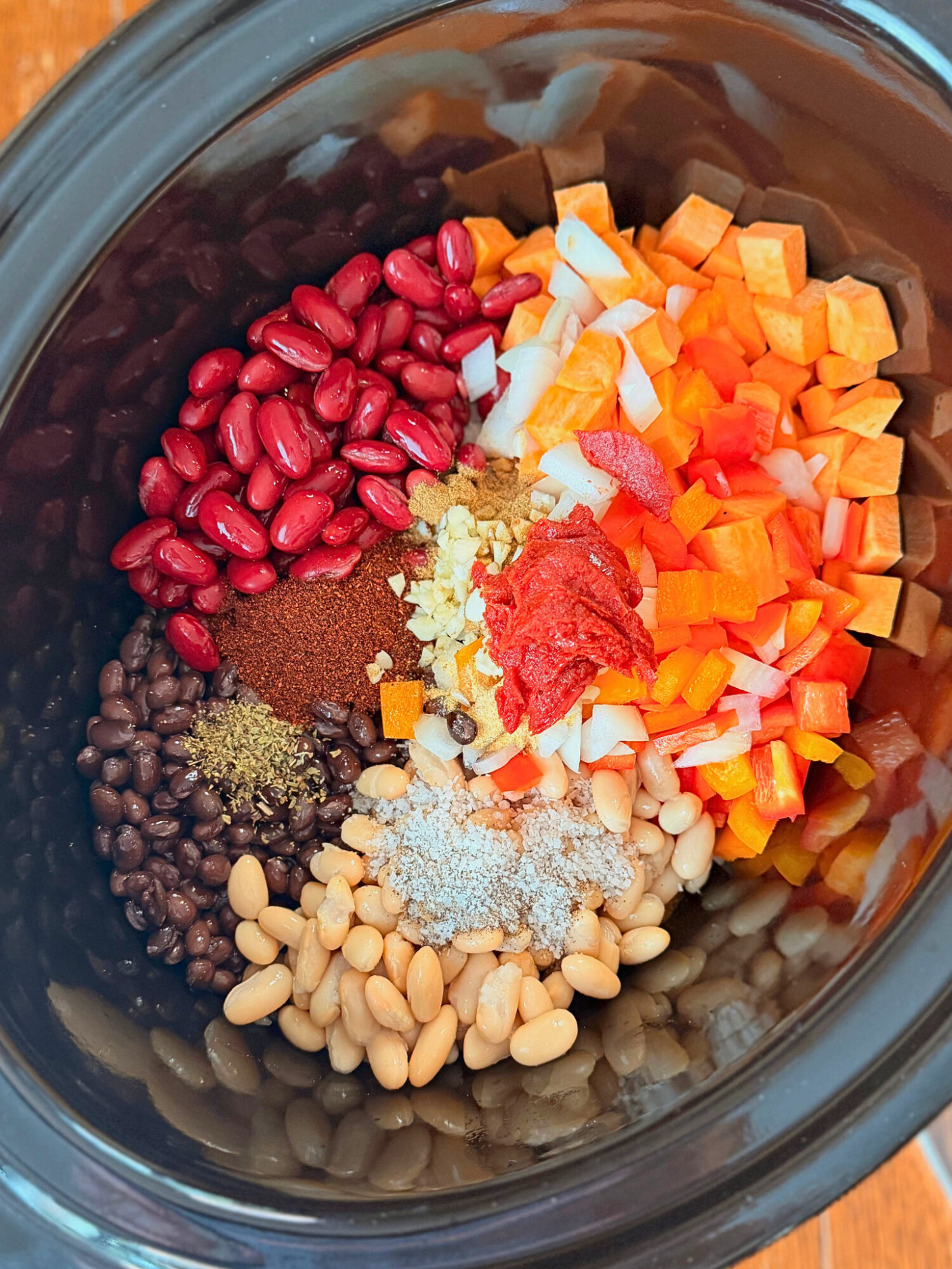 A slow cooker with three types of beans, spices, sweet potatoes, onions, and tomato paste.