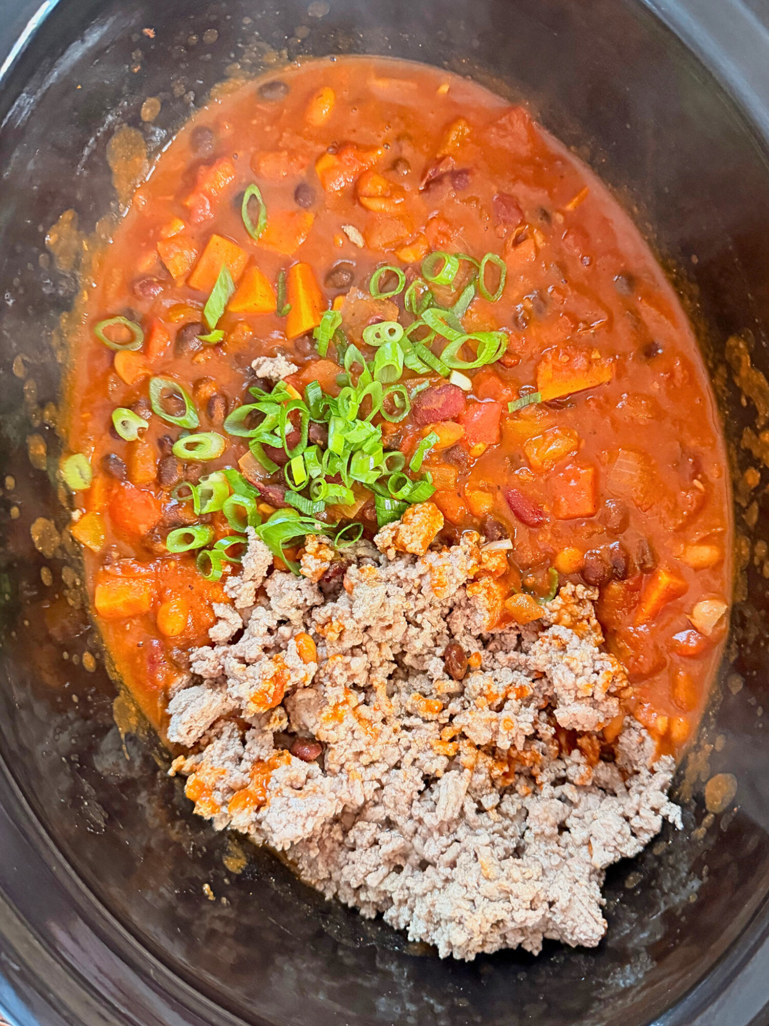 Adding ground turkey and chives to a slow cooker full of cooked chili.