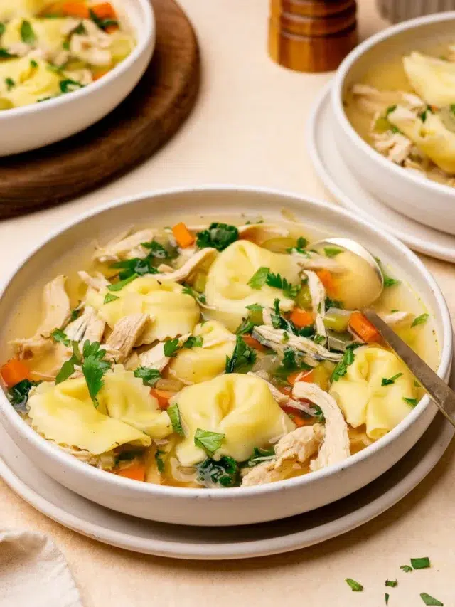 A bowl of chicken tortellini soup with two more bowls in the background.