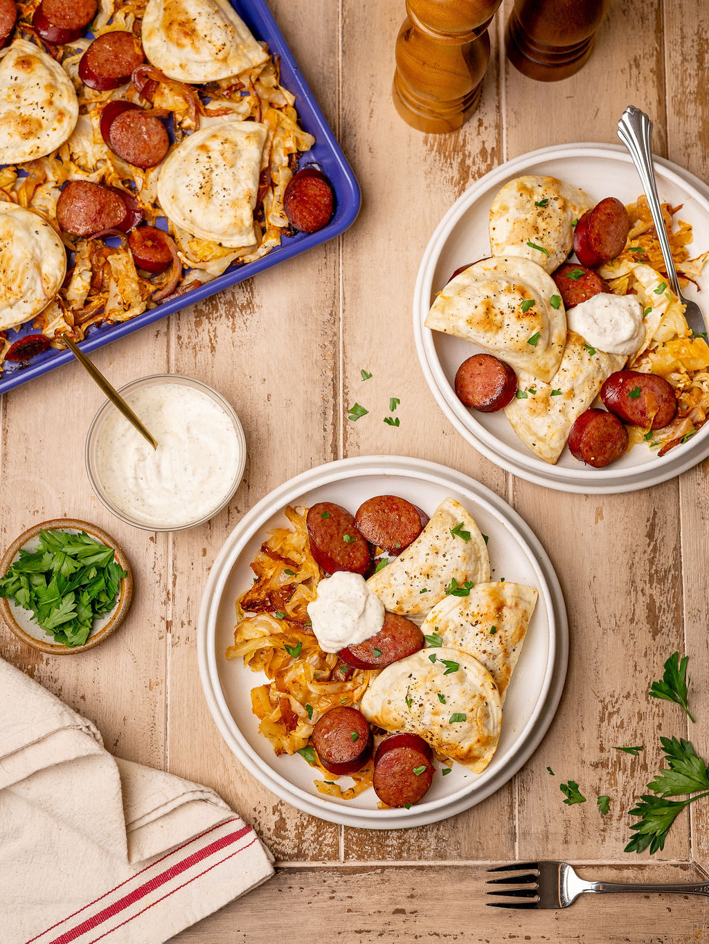 Two servings of pierogi and kielbasa sheet pan dinner over a light wooden surface surrounded by ingredients.