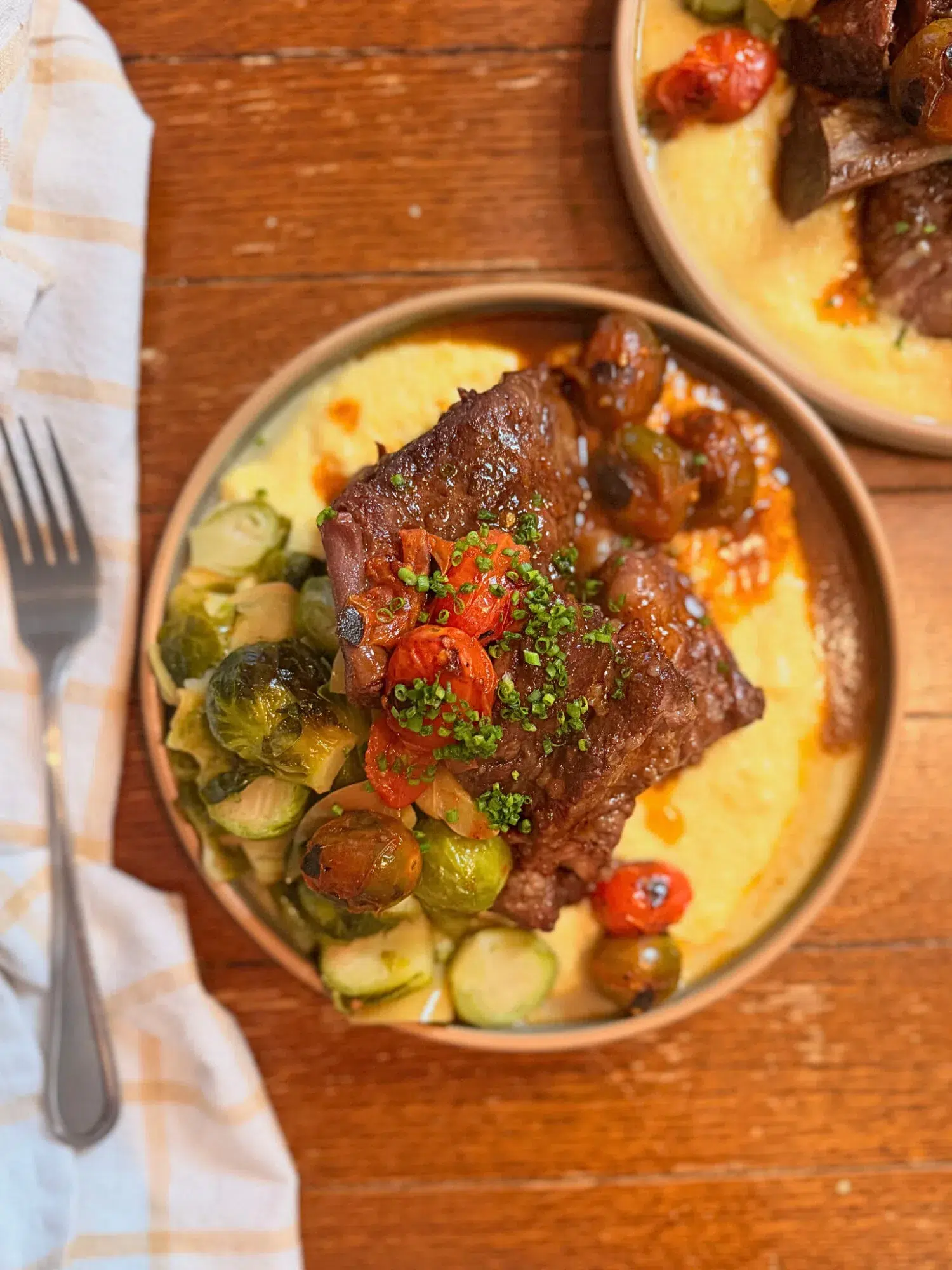 Short ribs in a bowl with polenta, cherry tomatoes, and brussels sprouts.