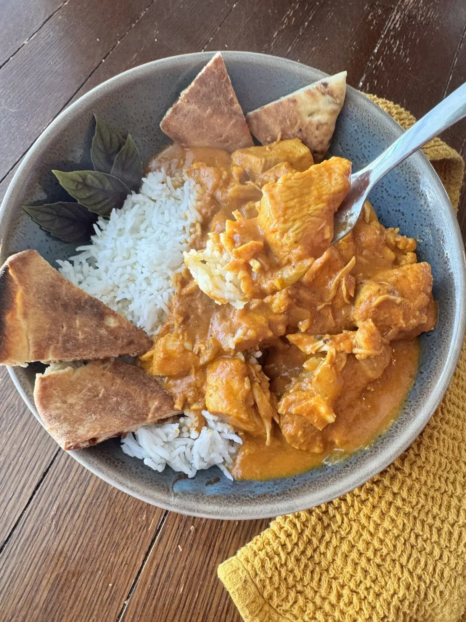 A fork dipping into a bowl of butter chicken over rice with naan.