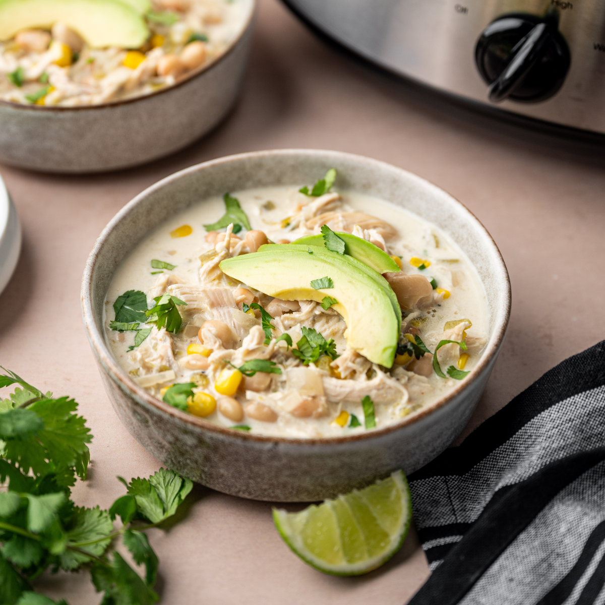 Slow cooker white chicken chili in a bowl topped with avocado and cilantro with slow cooker in background.