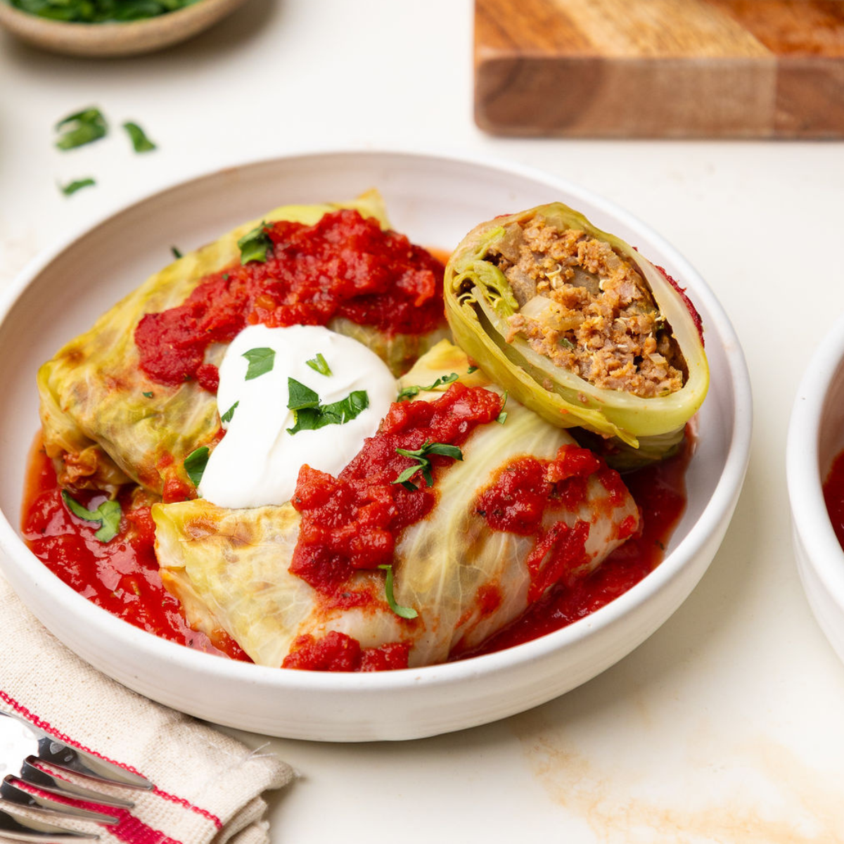 Lean beef stuffed cabbage rolls in a white bowl topped with tomato sauce and sour cream.