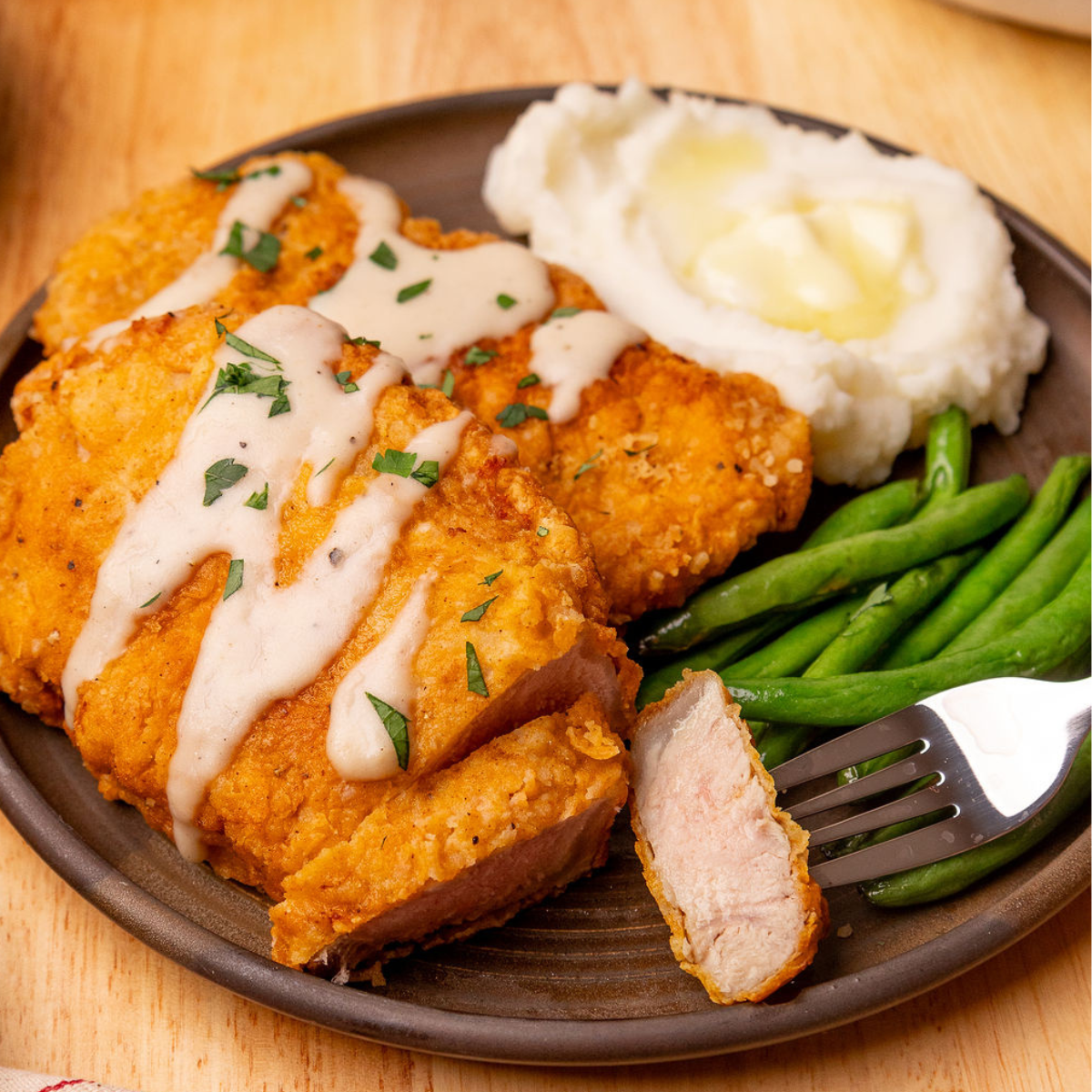 Taking a slice out of a chicken fried pork chop that's on a brown plate with a side of mashed potatoes and green beans.