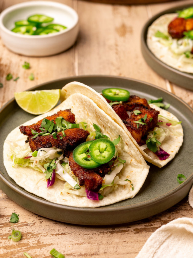 Two pork belly tacos on a plate with toppings.