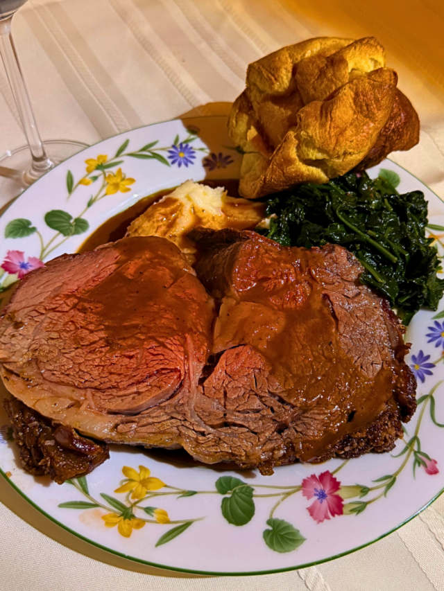 A slice of prime rib on a decorative plate served with sauted spinach and a dinner roll.