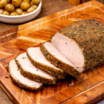 Herb crusted pork loin roast on na cutting board partially sliced with roasted potatoes in background.